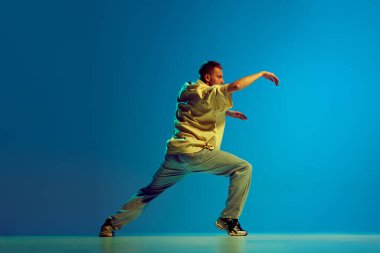 Şehir ritmi ve akışı. Gündelik kıyafetlerle dans eden yetenekli genç bir adamın dinamik görüntüsü neon ışıkta, değişken mavi arka planda hip-hop yapıyor. Sokak tarzı kavramı, modern dans, eğlence