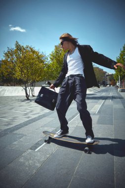 Siyah kostümlü, güneş gözlüklü, halka açık parkta hareket eden evrak çantalı kaykaylı şık bir genç. Eğlence ve özgürlük. Spor anlayışı, aktivite ve hareketler, hobi