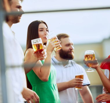 Bir grup arkadaş güneşli günün tadını çıkarır, soğuk biralarını yudumlarlar. Yeşil elbiseli kadın, gülümseyen yüzler ve gevşemiş titreşimlerle çevrili bir şekilde içki içer. Cuma havası, kutlama, Ekim Festivali kavramı.