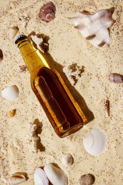 Cam bira şişesi kumlu sahilde, deniz kabuklarıyla çevrili, yaz tatili için ferahlatıcı bir içecek oluşturuyor. Markalaşma ve ürün yerleştirme reklamları, modelleme