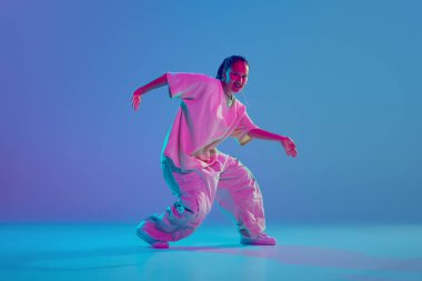 Aşırı büyük spor elbiseli genç artistik bir kızın neon rengi arka planda tutkulu bir şekilde hip hop dansının dinamik görüntüsü. Serbest stil dansı, modernlik, şehir kültürü, gençlik kavramı