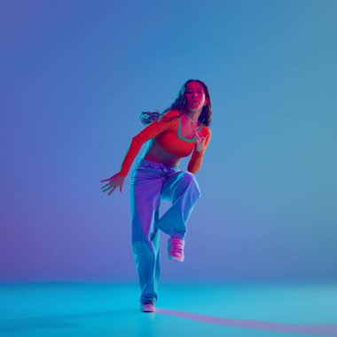 Enerji patlaması. Hareketli genç bir kız, neon ışıkta, değişken mavi arka planda, dinamik olarak hip hop yapıyor. Serbest stil dansı, modernlik, şehir kültürü, gençlik kavramı