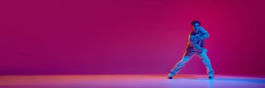 Hareketli gündelik giysiler içindeki enerjik genç adam, neon ışıkta pembe arka planda hip-hop dansı yapıyor. Serbest stil dansı, modernlik, şehir kültürü, gençlik kavramı. Pankart. İlan için alanı kopyala