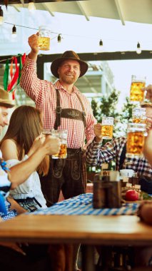 Kareli gömlek ve şapkalı neşeli sakallı adam, kutlama tostunda bira kadehi kaldıran neşeli adam, barda arkadaşlarıyla eğlenen. Ekim Festivali, festival, parti, bira fabrikası, gelenek kavramı