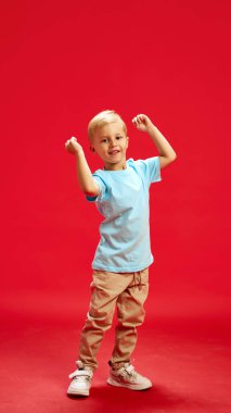Pozitif mutlu küçük çocuğun tam boy portresi, mavi tişörtlü ve kahverengi pantolonlu çocuk dans ediyor, kırmızı stüdyo arka planında eğleniyor. Çocukluk kavramı, duygular, moda, yaşam tarzı