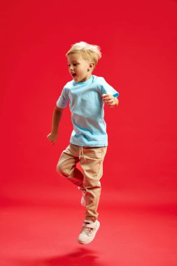 Mutlu küçük bir çocuğun tam boy görüntüsü, sarı saçlı günlük kıyafetler giymiş, kırmızı stüdyo arka planında oynuyor, zıplıyor. Çocukluk kavramı, duygular, moda, yaşam tarzı