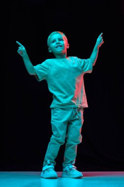 Genç bir çocuğun tam boy portresi, günlük kıyafetler içinde çocuk, kot pantolon ve tişört dansı, neon ışıklı siyah stüdyo arka planına karşı hareket halinde poz veriyor. Çocukluk, duygular, moda, yaşam tarzı konsepti