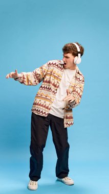 Sıradan giysiler içinde, kulaklıkla müzik dinleyen ve mavi stüdyo arka planında dans eden genç bir adamın tam boy portresi. İnsan duyguları, gençlik, yaşam tarzı, moda kavramı