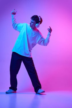 Gündelik giysiler içinde, kulaklıkla müzik dinleyen ve neon rengi pembe arka planda dans eden genç bir adamın tam boy portresi. İnsan duyguları, gençlik, yaşam tarzı kavramı