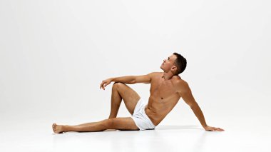 Yerde iç çamaşırlarıyla oturan yakışıklı bir adam, beyaz stüdyo arka planında yalnız poz veriyor. Gömleksiz, kaslı vücut. Erkek güzelliği, vücut ve cilt bakımı, sağlık ve spor anlayışı. Pankart