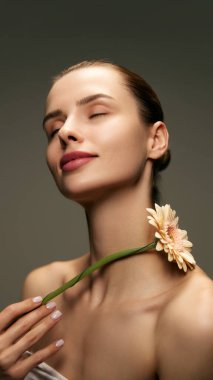 Pürüzsüz, pürüzsüz tenli, yeşil stüdyo arka planında Gerbera çiçeğiyle poz veren zarif genç bir kadının portresi. Doğal kadın güzelliği, cilt bakımı, kozmetik ve kozmetik bilimi, sağlık