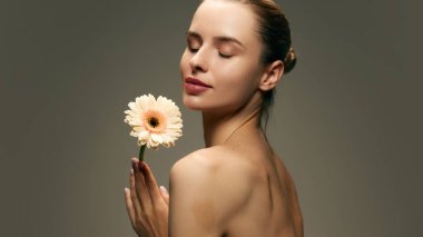 Pürüzsüz, lekesiz, yeşil stüdyo arka planında Gerbera çiçeğiyle poz veren güzel genç bir kadının portresi. Doğal kadın güzelliği, cilt bakımı, kozmetik ve kozmetik kavramı