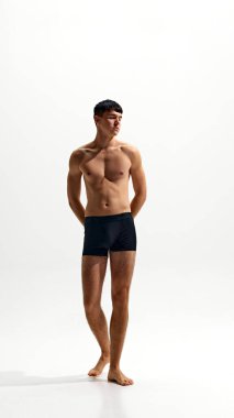 Beyaz stüdyo arka planında izole edilmiş siyah boksörlerle poz veren, rahatlamış, kaslı, yakışıklı genç bir adamın tam boy portresi. Erkek güzelliği, vücut ve cilt bakımı, sağlık ve spor kavramı