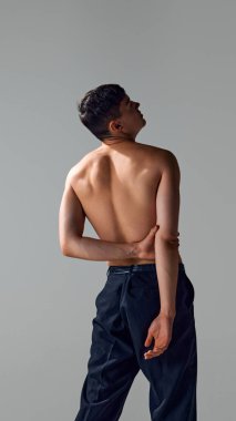 Gri stüdyo arka planında izole edilmiş pantolonla poz veren rahatlatıcı, güçlü, kaslı, sırt üstü çıplak bir adam. Erkek güzelliği, vücut ve cilt bakımı, sağlık ve spor kavramı