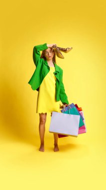 Dinamik alışveriş aktivitesinden sonra yorgun hissediyorum. Stüdyonun sarı arka planına karşı elinde bir sürü alışveriş çantasıyla duran genç bir kadın. Alışveriş, satış, Kara Cuma, Siber Pazartesi