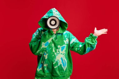 Yeşil kapüşonlu bir adam kırmızı stüdyo arka planına karşı megafonla konuşuyor. Haftalık Spesiyal Promosyon, teklifler. Satış, alışveriş, haber, duyuru, reklam kavramı