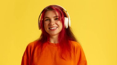 Turuncu kazak giyen, kulaklıkla müzik dinleyen ve sarı arka planda gülümseyen mutlu kızıl saçlı kadının portresi. İnsani duygular, aletler, rahatlama kavramı
