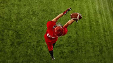 Yoğunlaşmış bir adamın en üst görüntüsü, kırmızı üniformalı Amerikan futbolcusu stadyumda hareket halinde, topu dikkatle yakalıyor. Spor anlayışı, rekabet, turnuva, oyun. Boşluğu kopyala