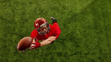 Yoğunlaşmış bir adamın en üst görüntüsü, kırmızı üniformalı Amerikan futbolcusu stadyumda hareket halinde, topu dikkatle yakalıyor. Spor anlayışı, rekabet, turnuva, oyun. Boşluğu kopyala