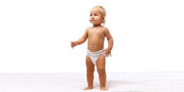 Merak ve sükunet. Güzel küçük bir kız bebek, bebek bezi takmış, sakin bir ifadeyle yatakta duruyor, beyaz stüdyo arka planında soyutlanmış. Çocukluk kavramı, annelik, bakım