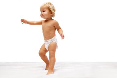İlk adımları atıyorum. Güzel küçük bir kız bebek, bebek bezi içinde ayakta duruyor ve beyaz stüdyo arka planında tek başına yürümeye çalışıyor. Çocukluk kavramı, annelik, bakım