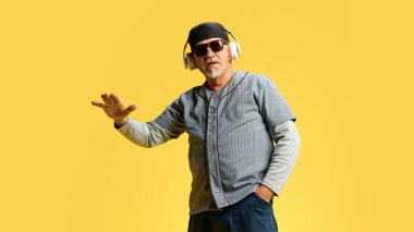 Şapkalı ve güneş gözlüklü yaşlı adam sarı stüdyo arka planında kulaklıkla müzik dinliyor. Şık bir DJ. Keyif. İnsan duyguları kavramı, yaşlı yaşam tarzı. Boşluğu kopyala, ad