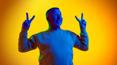 Mutlu, mutlu yüzlü yaşlı bir adamın portresi, barış jesti gösteriyor ve neon ışıkta sarı arka plana gülümsüyor. İnsan duyguları kavramı, yaşlı yaşam tarzı. Boşluğu kopyala, ad