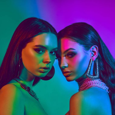 Moda makyajlı iki zarif genç kadının yakın plan portresi, parlak arka planda kalın küpeler. Kadın güzelliği, cazibe ve modern moda kavramı