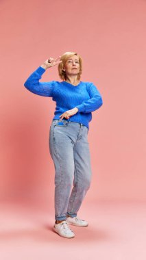 Gülümseyen, mutlu, olgun, günlük kıyafetler içinde, kot pantolon ve kazak giyen, pembe stüdyo arka planında dans eden yaşlı bir kadının tam boy portresi. Yaşlıların yaşam tarzı, duyguları, bakımı, sağlığı kavramı