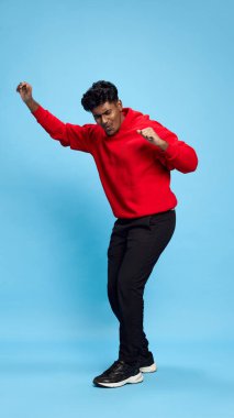 Siyah kot pantolon ve kırmızı kapüşonlu neşeli Hintli genç bir adamın tam boy portresi mavi stüdyo arka planında neşeli dans ediyor. Başarıyı kutluyoruz. Duyguların kavramı, yaşam tarzı