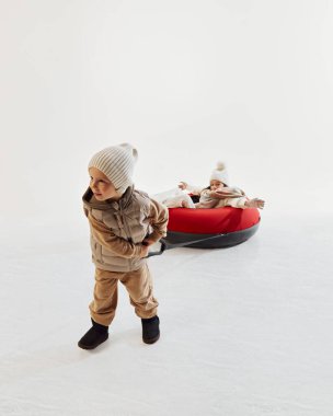 Kış kıyafetleri içinde küçük bir çocuk, kız kardeşini kırmızı kar tüpüne çekiyor, kış boyunca aile aktivitelerinin sıcaklığını ve neşesini gösteriyor. Mutluluk. Kış mevsimi kavramı, aktiviteler, eğlence, eğlence