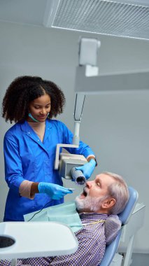 Diş hekimi detaylı diş röntgeni çekmeye, hastaya süreç boyunca rehberlik etmeye ve en iyi görüntüleme sonuçları için rahat kalmasını sağlamaya hazırlanıyor. Sağlık sigortası, güzellik, hijyen, dişçi kavramı. Ad.