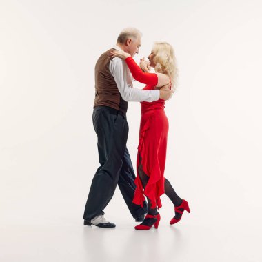 Elderly woman and senior man, elegantly dressed in red dress and black suit, dancing tango with passion isolated on white background. Concept of retro dance, hobby, aging, active lifestyle clipart