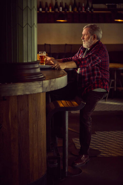 Пожилой бородатый мужчина в клетчатой рубашке сидит в тёплом освещенном баре, пьёт пиво, разговаривает с барменом и отдыхает. Концепция образа жизни пожилых людей, напитков, ретро-моды