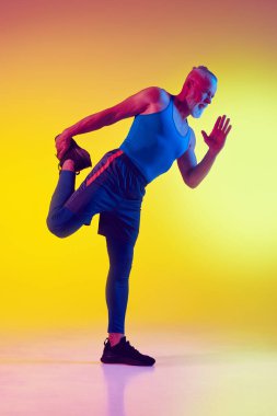 Spor giyim sektöründe sportif yaşlı bir adam tek ayak üzerinde dengede duruyor, antrenman yapıyor, neon renkli sarı arka plana karşı atletik vücudunu gösteriyor. Spor anlayışı, aktif yaşlı yaşam tarzı, sağlık hizmetleri