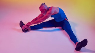 Kas gücü kuvvetli vücudu olan yaşlı bir adam spor giyimi eğitiminde, neon ışıkta eğimli sarı arka plana karşı esneme egzersizleri yapıyor. Spor anlayışı, aktif yaşlı yaşam tarzı, sağlık hizmetleri