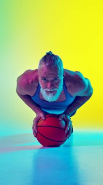 Spor kıyafetli yaşlı kaslı bir adam, basketbol topunun üzerinde, neon ışıkta yeşil arka planda duran gradyan sarı renge karşı duruyor. Ön manzara. Spor anlayışı, aktif yaşlı yaşam tarzı, sağlık hizmetleri