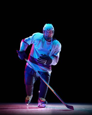 Rekabetçi erkeklerin dinamik görüntüsü, beyaz spor giyimli hokey oyuncusu sopayla ringde hareket ediyor, neon ışıkta siyah stüdyonun arka planına karşı. Spor, oyun, turnuva, yarışma kavramı