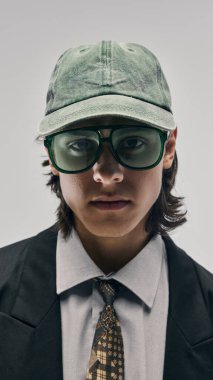 Yeşil şapkalı, büyük yeşil gözlüklü, siyah ceketli, beyaz gömlekli ve stüdyo arka planında poz veren şık bir gencin yakın plan portresi. Moda anlayışı, şehir tarzı, reklam