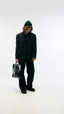 Moda ileri bakışı. Şapkalı, siyah kostümlü, gözlüklü, deri çantalı beyaz stüdyo arka planında poz veren şık bir gencin tam boy portresi. Moda anlayışı, şehir tarzı, reklam