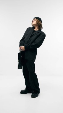 Bereli, siyah kostümlü, gözlüklü, deri çantalı, beyaz stüdyo arka planında poz veren kendinden emin bir gencin tam boy portresi. Moda kavramı, gençlik kültürü, şehir tarzı, reklam