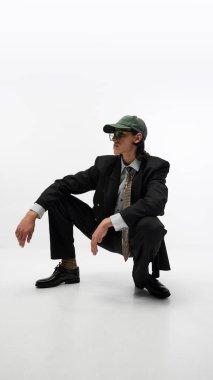 Gündelik ve resmi tarzda giyinen genç adam, desenli kravat, siyah ceket, yeşil başlık ve yeşil güneş gözlüğü takıyor, beyaz stüdyo arka planında poz veriyor. Moda kavramı, gençlik kültürü, reklam