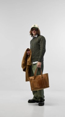 Portrait of stylish young man in green sweater over collared shirt, paired with green trousers and beige cap, posing with leather tote bag. Concept of fashion, youth culture, urban style, ad clipart