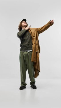 Şık yeşil örgü süveter giyen yakışıklı genç adam, uyumlu pantolon, bej şapka, trençkot giyiyor, beyaz stüdyo arka planında poz veriyor. Moda kavramı, gençlik kültürü, şehir tarzı, reklam