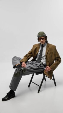 Yakışıklı ve kendine güvenen genç adam sandalyede poz veriyor, eski püskü ceket giyiyor, gri pantolon giyiyor, büyük boy gözlük ve şapka takıyor. Retro-modern titreşim. Moda, gençlik kültürü, şehir tarzı, reklam konsepti.