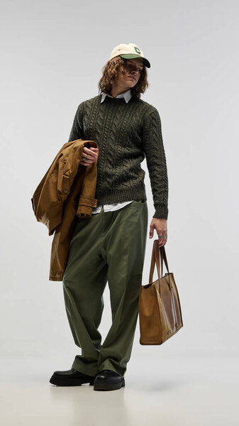 Стильный и красивый молодой человек в зеленом свитере поверх воротничковой рубашки, в паре с зелеными брюками и бежевой кепкой, позируя с кожаной сумкой тотализатора и плащом. Концепция моды, молодежи, городского стиля, объявления