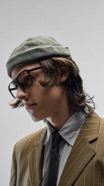 Moda şapkalı, büyük boy gözlüklü, klasik kravatlı çizgili ceketli beyaz stüdyo arka planında poz veren yakışıklı bir gencin yakın plan portresi. Moda kavramı, gençlik kültürü, şehir tarzı, reklam