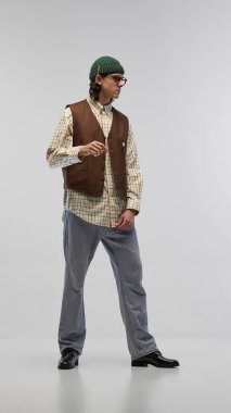 Retro öğrenci tarzı. İşaretli gömlekli, kahverengi yelekli, kot pantolonlu, yeşil bereli ve gözlüklü beyaz stüdyo arka planında poz veren yakışıklı bir delikanlı. Moda anlayışı, gençlik, şehir tarzı, reklam