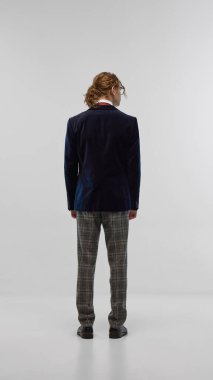 Lacivert kadife ceketli, damalı gri pantolonlu ve klasik siyah ayakkabılı beyaz stüdyo arka planında poz veren genç bir adam. Moda, gençlik kültürü, zarif tarz, reklam konsepti.