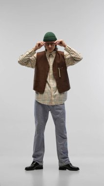 Tam boy. Yakışıklı, şık bir genç adam. İşaretli gömlek, kahverengi yelek, yeşil bereli günlük kot pantolon ve stüdyo arka planında gözlükler. Moda anlayışı, gençlik, şehir tarzı, reklam